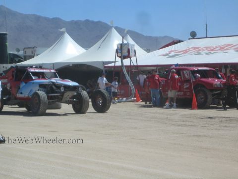 1990 Ford Bronco - Off-road race trucks stage on a dusty course beside white peaked tents and a red pit canopy as crew and spectators stand around.