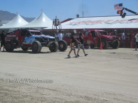 1990 Ford Bronco - Off-road race trucks sit at the red Terrible Herbst pit as spectators walk past, with an American flag and camera crane overhead.
