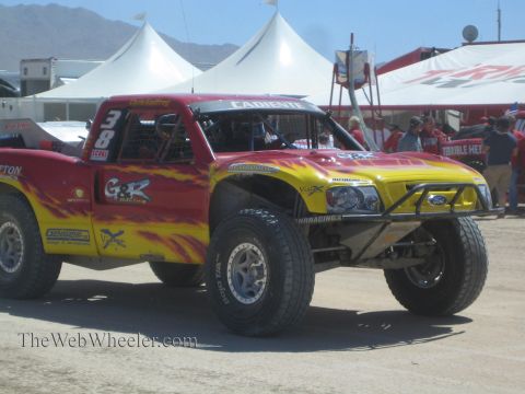1990 Ford Bronco - red and yellow flame-painted Ford off-road race truck numbered 38 in G&R Racing livery sits in a dusty pit beside white tents.