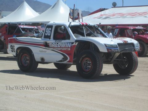 1990 Ford Bronco - white and red Baldwin Motorsports off-road race truck numbered 96 sits in a dusty pit area beside white tents and other race trucks.