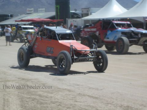 1990 Ford Bronco - orange tube-frame off-road race buggy speeds across a dusty course past other staged race trucks and spectators.
