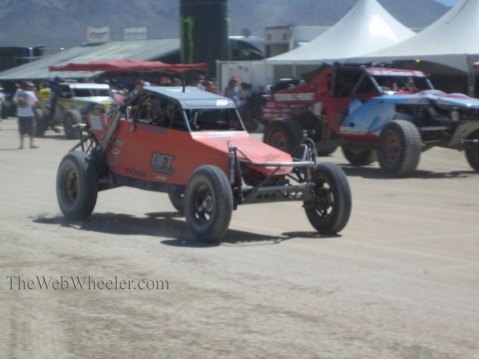 Orange tube-frame off-road buggy racing on dirt past other race trucks