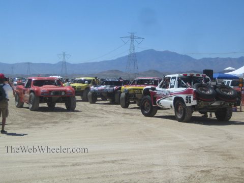 1990 Ford Bronco - row of off-road race trucks, including a white and red number 96, stages on a dusty desert course beneath power-line towers and mountains.