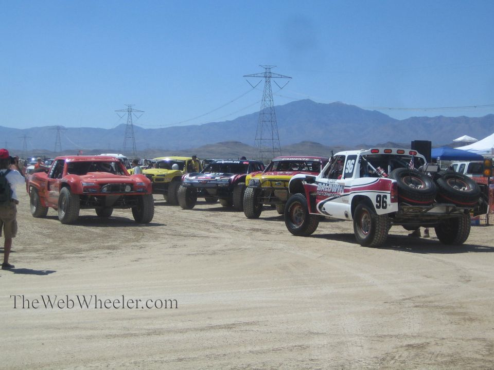 Row of off-road race trucks including number 96 staged on dirt with power-line towers