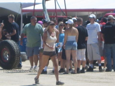 1990 Ford Bronco - crowd of spectators gathers at an off-road racing event beside coolers and a race-truck tire as a woman in shorts walks in the foreground.