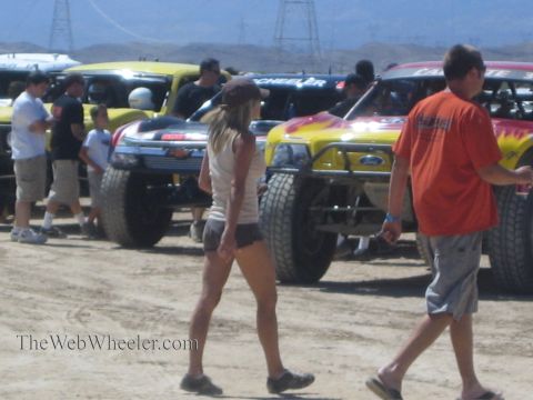 1990 Ford Bronco - Spectators walk past a line of staged off-road race trucks, including yellow and flame-painted Ford trucks, on a dusty desert course.
