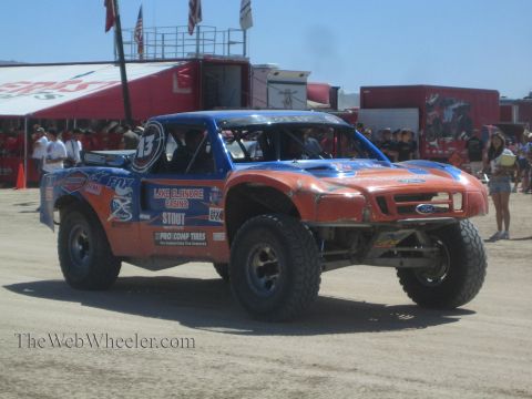 1990 Ford Bronco - blue and orange Lake Elsinore Casino Ford off-road race truck numbered 13 drives through a dusty pit area past spectators.