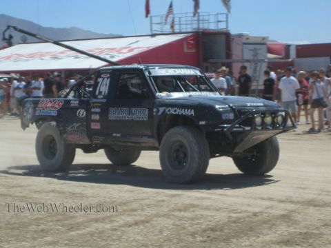 1990 Ford Bronco - black Board Ford Motorsports off-road race truck numbered 741 in Lucas Oil and Yokohama livery drives through a dusty pit past spectators.