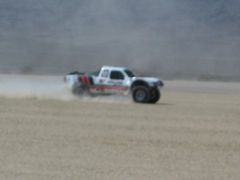 1990 Ford Bronco - white off-road race truck races across a desert dry lakebed, trailing a plume of dust with mountains behind.