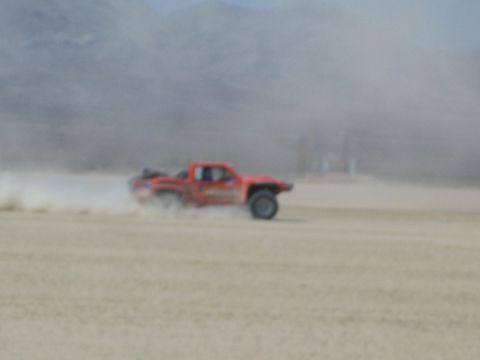 1990 Ford Bronco - orange off-road race truck speeds across a dusty desert flat, kicking up a trail of dust under a hazy sky.