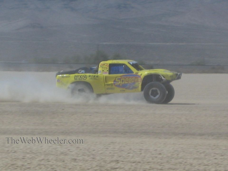 Yellow Speedco off-road race truck drifting across desert lakebed in dust