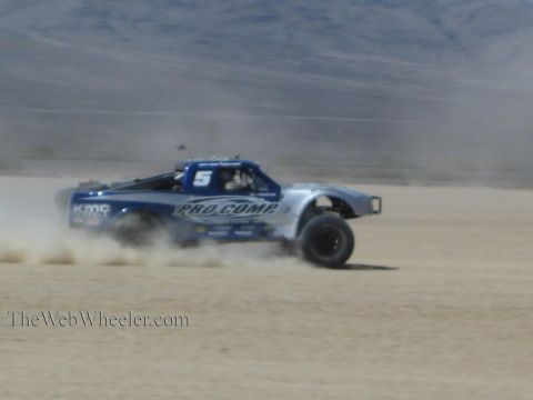 1990 Ford Bronco - blue and silver off-road race truck numbered 5 with Pro Comp livery races across a dusty desert flat.
