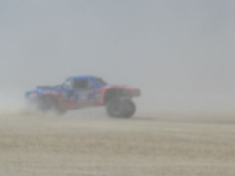 1990 Ford Bronco - red and blue off-road race truck powers through thick dust on a hazy desert flat.