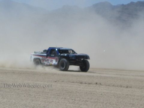 1990 Ford Bronco - blue off-road race truck with Lucas Oil livery powers through heavy dust with mountains in the background.