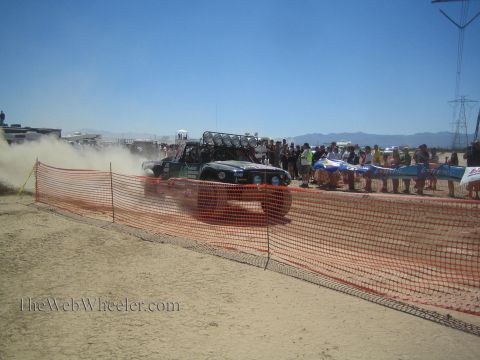 1990 Ford Bronco - off-road vehicle kicks up dust passing a crowd of spectators behind an orange safety fence at a desert race.