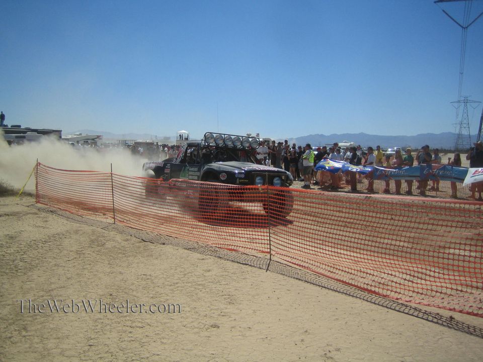 Off-road vehicle passing spectators behind orange fence at desert race