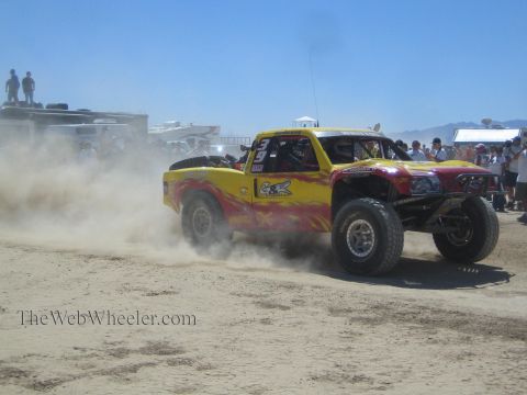 1990 Ford Bronco - yellow off-road race truck with red flames launches from the start area, spraying sand before a crowd.