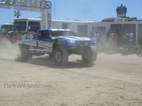 1990 Ford Bronco - blue Jimco off-road race truck numbered 20 launches from a desert race start under a banner with spectators watching.