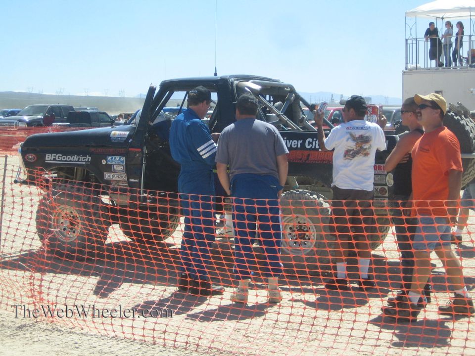 People standing around black race truck behind orange fence in pit
