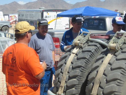 1990 Ford Bronco - Several men in caps and racing suits talk beside two large off-road tires at a dusty desert race pit.