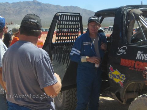 1990 Ford Bronco - driver in a blue racing suit stands by a black off-road truck talking with crew members in a desert pit.