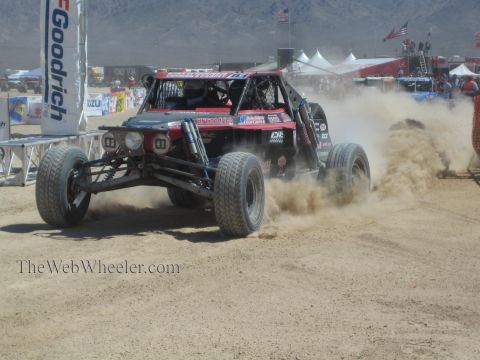 1990 Ford Bronco - red and black open-wheel off-road race buggy sprays sand as it races past banners at a desert event.