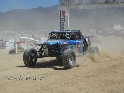 1990 Ford Bronco - blue open-wheel off-road race buggy numbered 115 kicks up sand racing under a BFGoodrich banner in the desert.