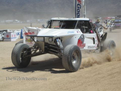 1990 Ford Bronco - white open-wheel off-road race buggy speeds across sand as a marshal waves a checkered flag.