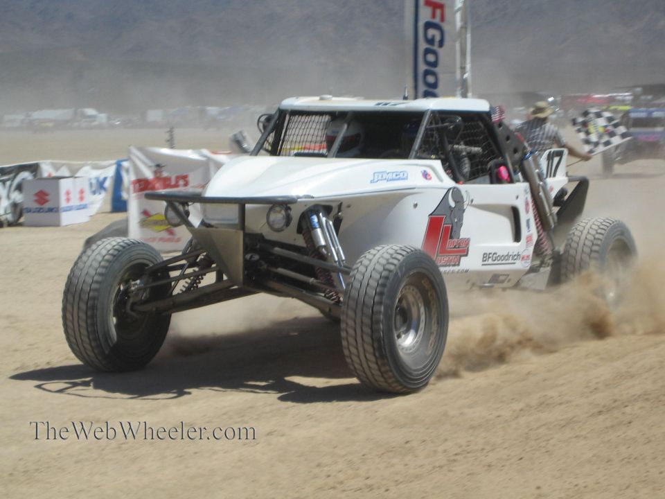White open-wheel off-road buggy racing past a checkered flag in desert
