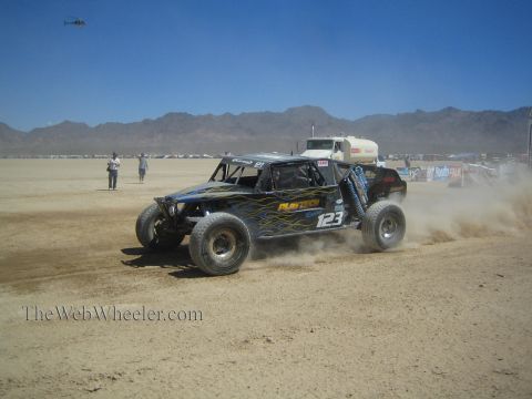 1990 Ford Bronco - dark open-wheel off-road race buggy numbered 123 races across the desert as a helicopter hovers overhead.