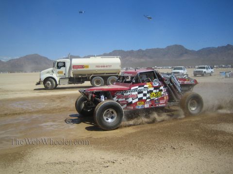 1990 Ford Bronco - red open-wheel off-road race buggy numbered 124 sprays dirt racing past a parked fuel tanker truck in the desert.