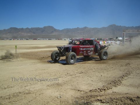 1990 Ford Bronco - maroon tube-chassis off-road race buggy marked 126 races across a dusty desert dry-lake course with mountains behind.