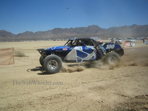 1990 Ford Bronco - blue and white off-road race buggy numbered 100 with McMillin Homes livery slides through desert dirt near orange fencing.