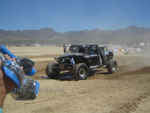 1990 Ford Bronco - black off-road race buggy runs a desert dry-lake course while a spectator with a blue wrapped item stands in the foreground.