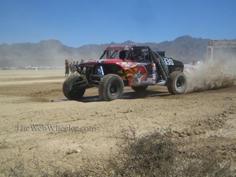 1990 Ford Bronco - dark red off-road race buggy numbered 30 with flame graphics throws a dirt rooster tail on a desert race course.