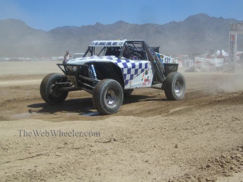 1990 Ford Bronco - blue and white checkered off-road race buggy numbered 131 corners hard, kicking up dust on a desert course.