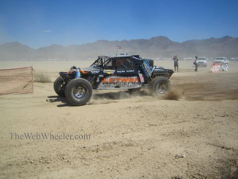 1990 Ford Bronco - black off-road race buggy with Letner livery throws up dirt while racing a desert dry-lake course past photographers.