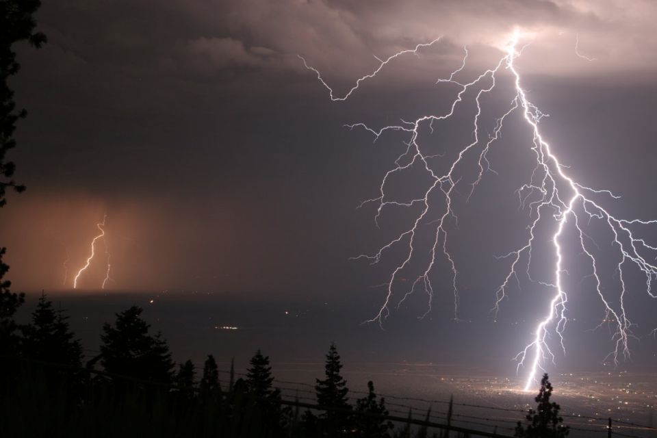 Lightning strikes illuminate the sky over a distant city at night, with trees in the foreground