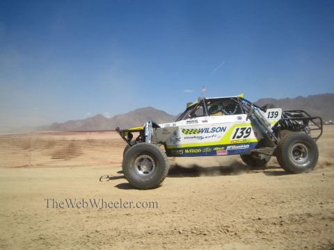 1990 Ford Bronco - white and green off-road race buggy numbered 139 with Wilson Motorsports livery races a desert course beside orange fencing.