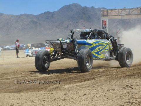 1990 Ford Bronco - silver, blue and yellow open tube-chassis off-road buggy races across a desert course as a spectator watches.