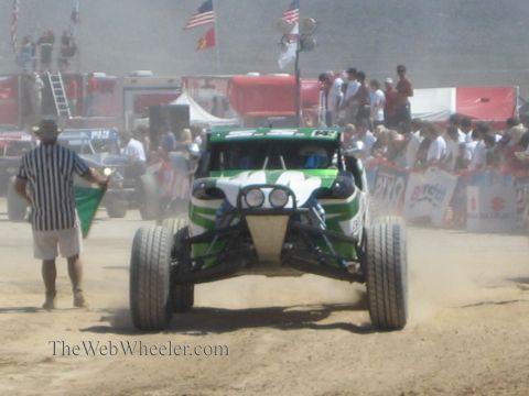 1990 Ford Bronco - green and white off-road race buggy approaches head-on past a striped-shirt flagman and a crowd lining the desert course.