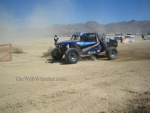 1990 Ford Bronco - blue and white off-road race buggy slides through dirt past orange fencing on a desert dry-lake course.