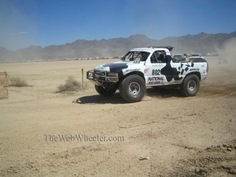 1990 Ford Bronco - white off-road race truck numbered 802 with cow-print and checkered graphics races a desert course.