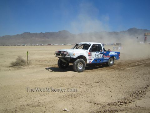 1990 Ford Bronco - white and blue off-road race pickup with Voss livery corners through dust on a desert race course.