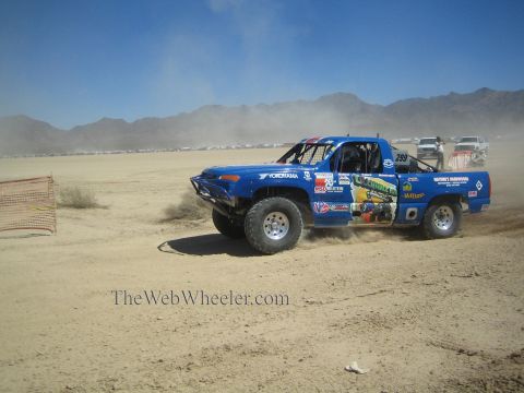 1990 Ford Bronco - blue off-road race pickup with Yokohama livery corners through dirt past orange fencing on a desert course.