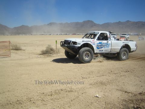 1990 Ford Bronco - white #701 off-road race truck throws up dust as it races across a flat desert course backed by distant mountains.