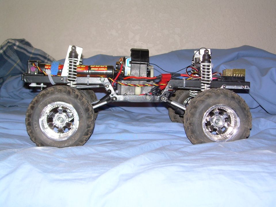 Side profile of an RC rock crawler chassis with chrome wheels and red coilovers on a bed