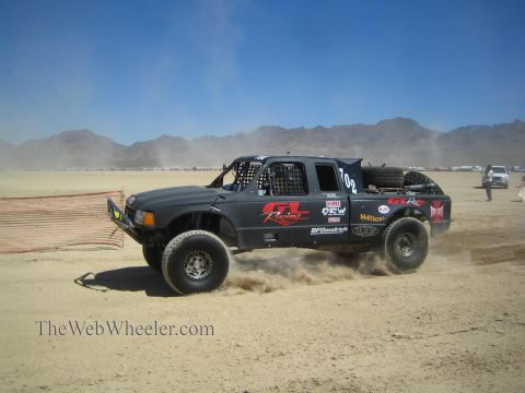 1990 Ford Bronco - black off-road race truck marked GR Racing and numbered 102 speeds across a dusty desert course near mountains.