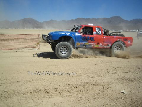 1990 Ford Bronco - red-and-blue off-road race truck numbered 703 slides through the dust on a flat desert race course.