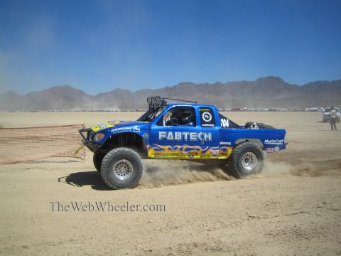 1990 Ford Bronco - blue Fabtech-liveried off-road race truck numbered 704 races through dust on a desert course backed by mountains.